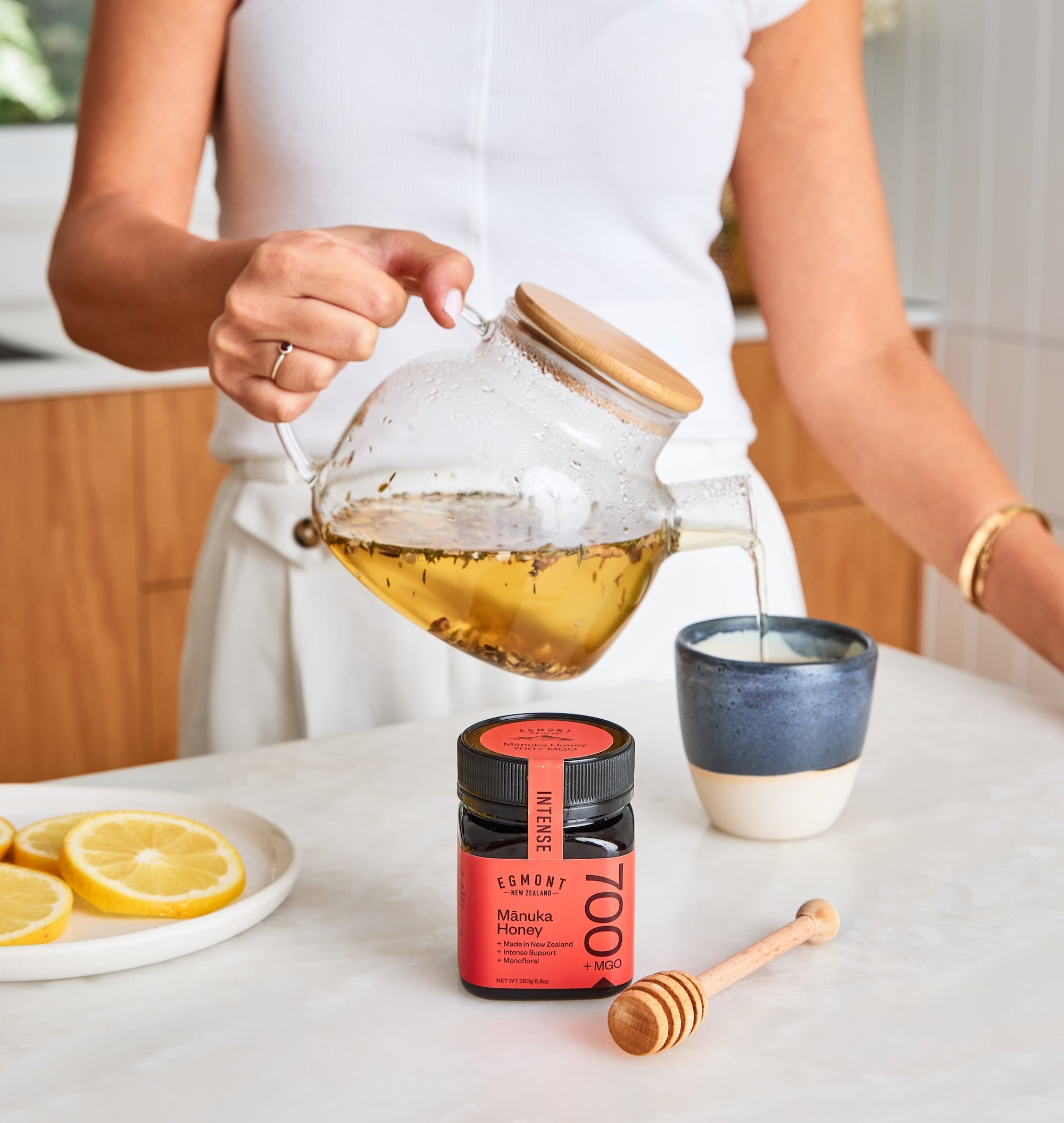 Person pouring tea into a cup with a jar of honey and lemon slices on a table.